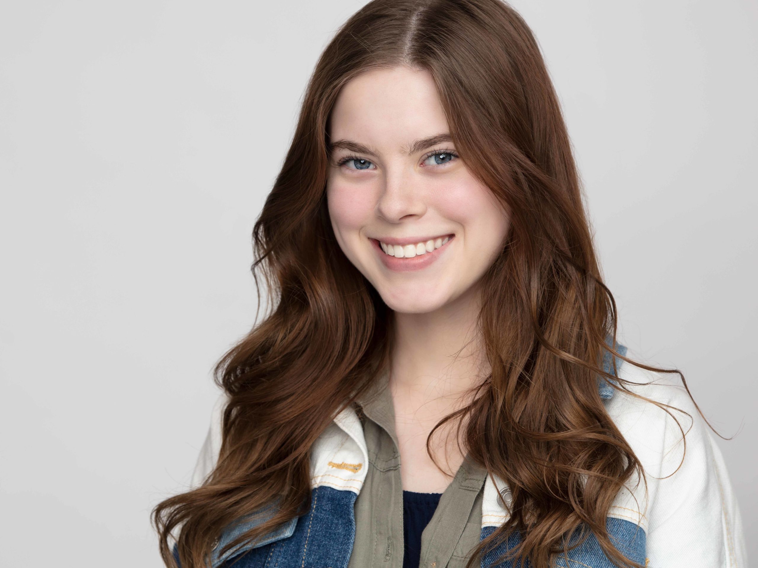 Actor headshot of a young brunette woman wearing a blue and white jean jacket over a brown button-up shirt, photographed against a gray background for a Dallas portfolio session