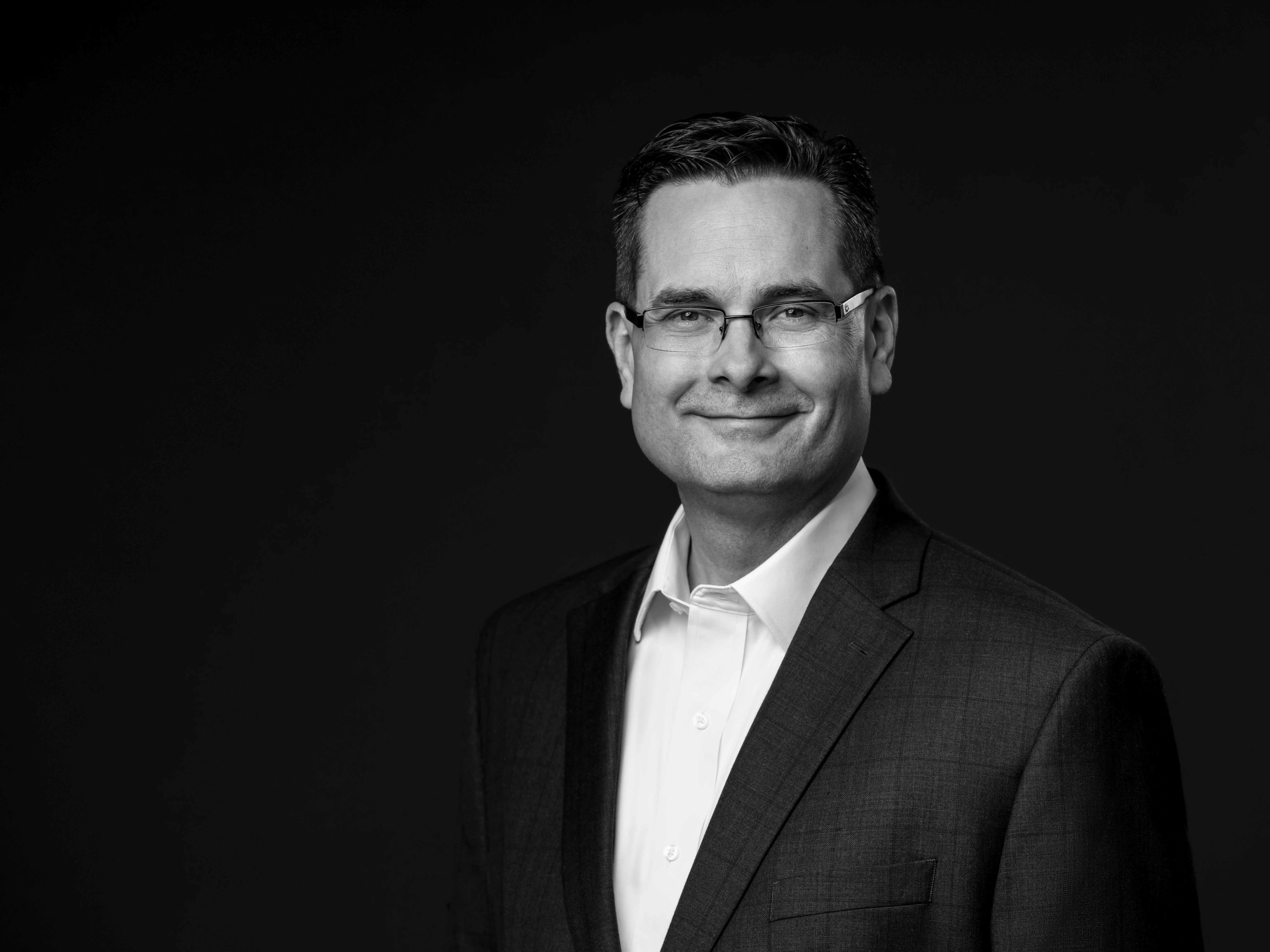 Black and white professional headshot of a young adult man in a suit, photographed against a plain black background