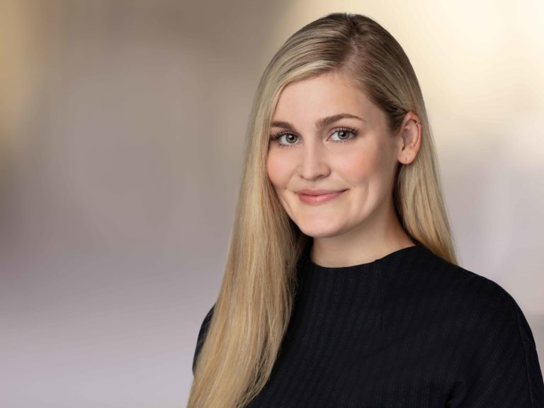Professional headshot of a young blonde woman wearing a black top, photographed against a soft, blurred environmental background