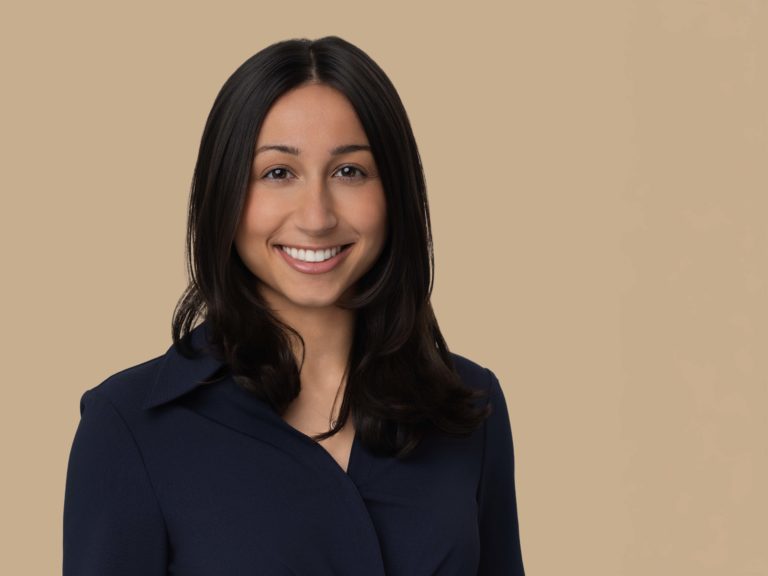 Professional headshot of a young woman in a blue blouse against a tan background